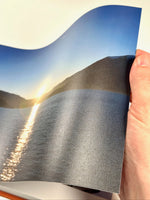 Hoja del fotolibro sostenida en la mano con una foto de atardecer sobre el lago, mostrando la flexibilidad del papel y la apertura plana