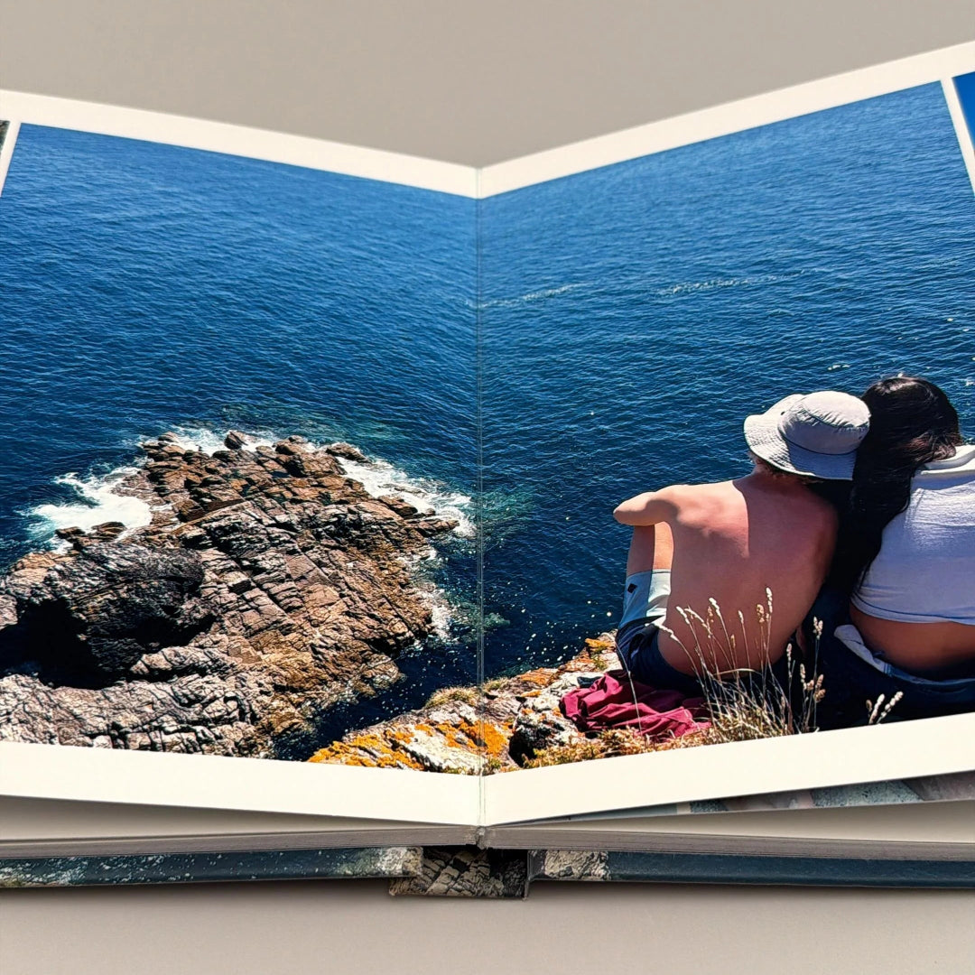 Fotolibro abierto a doble página con apertura 180°, mostrando una pareja frente al mar entre rocas de la costa