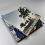 Dos fotolibros apilados con tapa fotográfica de playa y palmeras, mostrando el grosor y el acabado laminado mate
