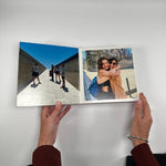 Manos sosteniendo un fotolibro abierto a doble página con fotos de viaje impresas en papel fotográfico