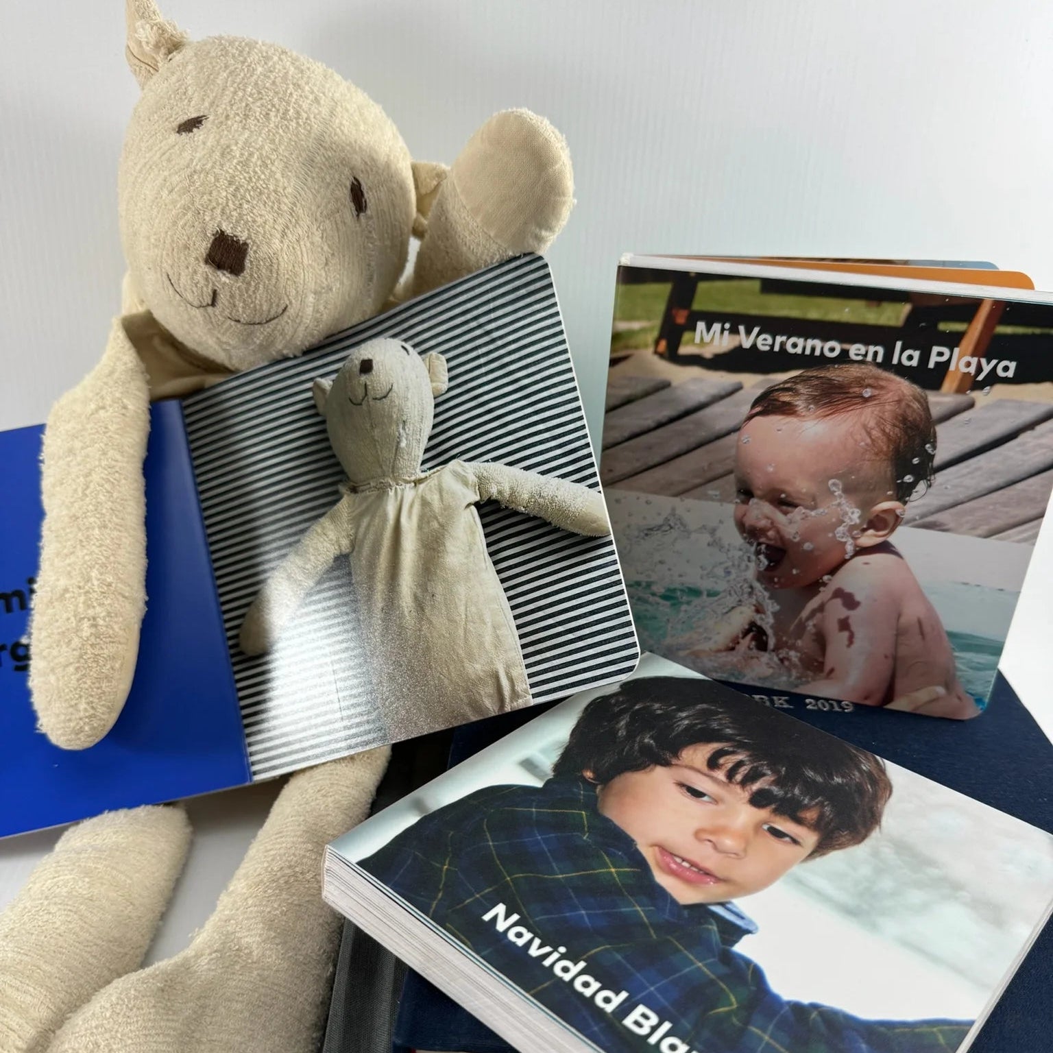 Oso de peluche sosteniendo un pequeño fotolibro a rayas junto a otros álbumes con fotos de bebés y niños