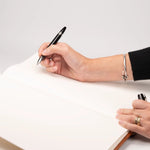 Persona escribiendo con lapicera negra sobre las páginas ahuesadas en blanco del libro de visitas abierto