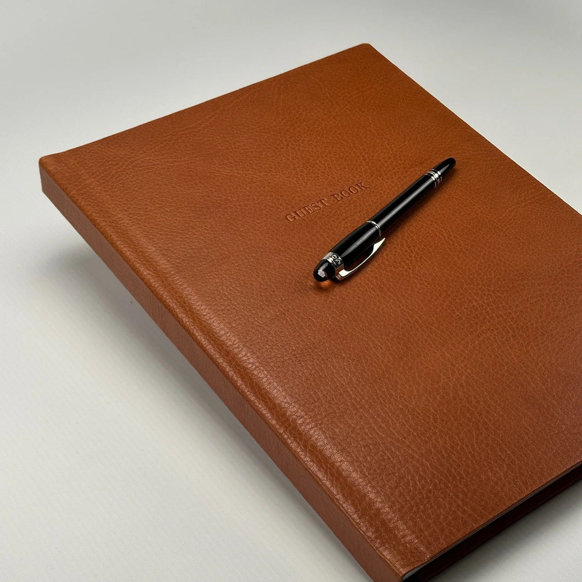 Libro de firmas de cuero color cognac cerrado, con la leyenda Guest Book grabada y una lapicera apoyada encima