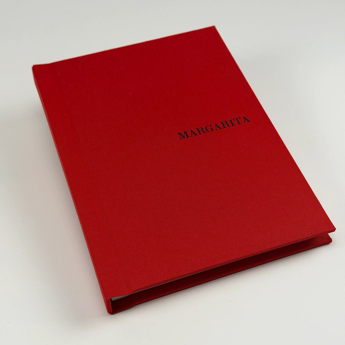 Cuaderno de lino rojo personalizado con el nombre MARGARITA grabado en negro, visto en ángulo sobre fondo claro