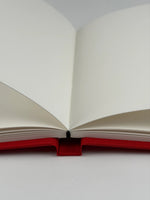 Cuaderno abierto a doble página mostrando el papel ahuesado texturado y la encuadernación cosida a mano sobre tapa de lino rojo