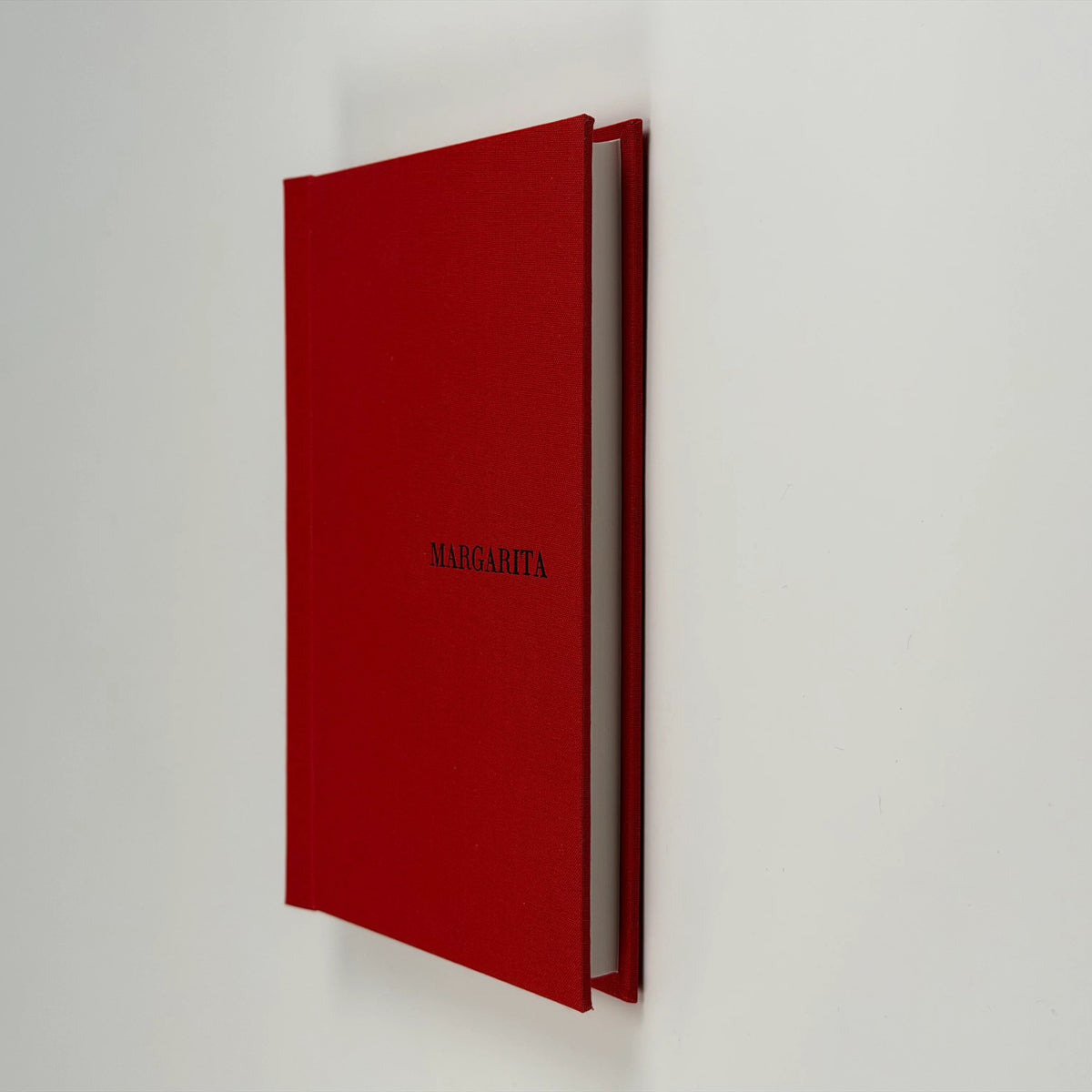 Cuaderno de tapa de lino rojo apoyado de canto y levemente abierto, con el nombre MARGARITA grabado en la tapa