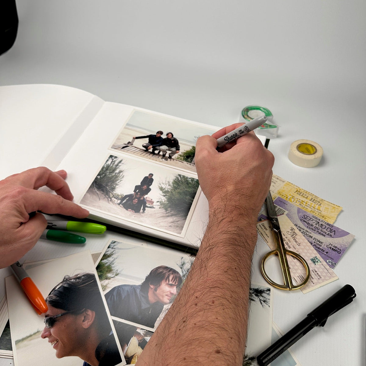 Mano escribiendo con fibrón sobre las páginas del álbum, rodeado de fotos impresas, tijera y tickets