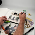 Mano escribiendo con fibrón sobre las páginas del álbum, rodeado de fotos impresas, tijera y tickets