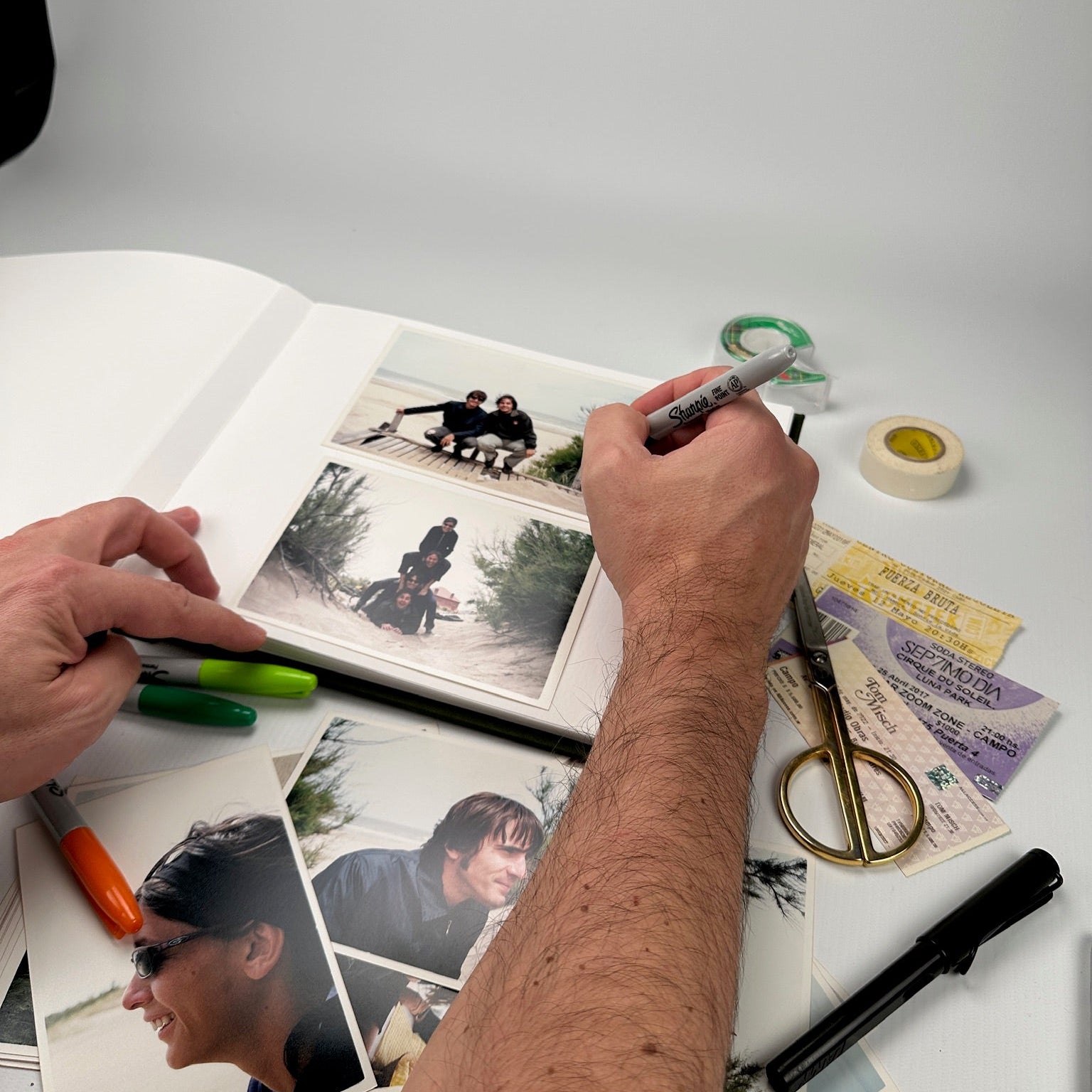 Mano escribiendo con fibrón sobre las páginas del álbum, rodeado de fotos impresas, tijera y tickets