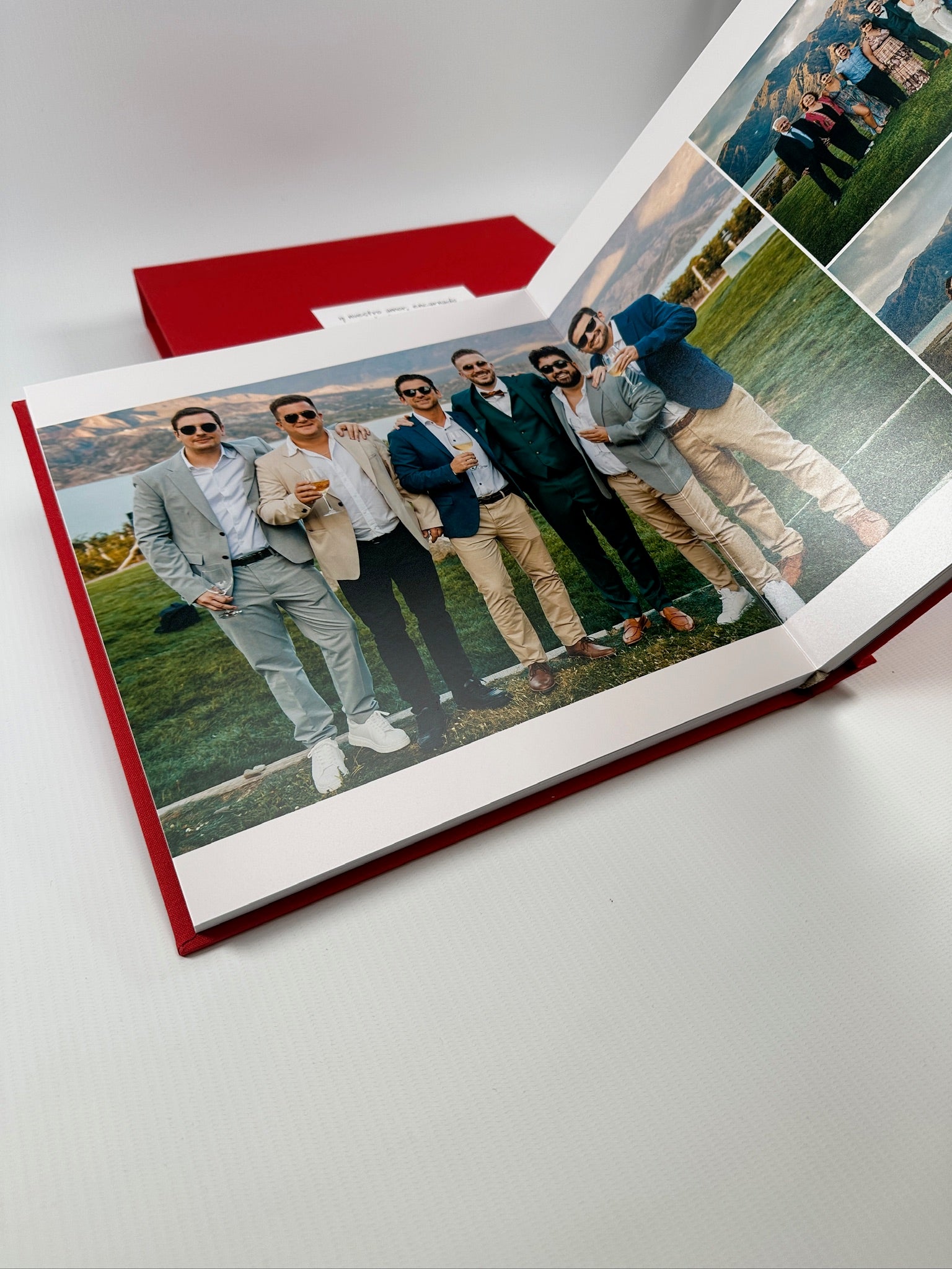 Álbum de boda abierto con foto grupal a página completa, apoyado sobre el estuche rojo