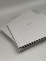 Fotolibro de lino gris claro con nombre en plateado, apoyado sobre su caja a juego