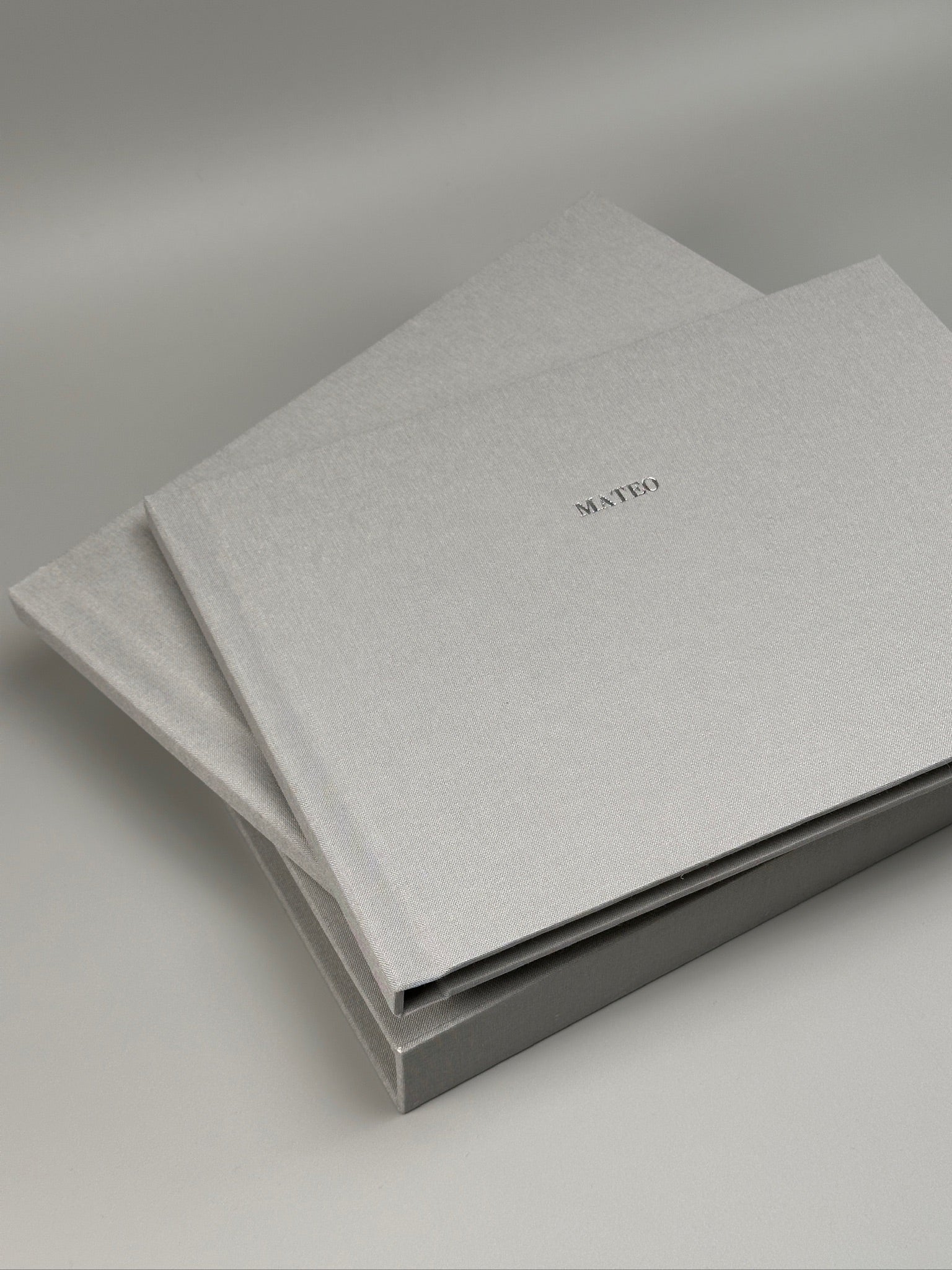 Fotolibro de lino gris claro con nombre en plateado, apoyado sobre su caja a juego