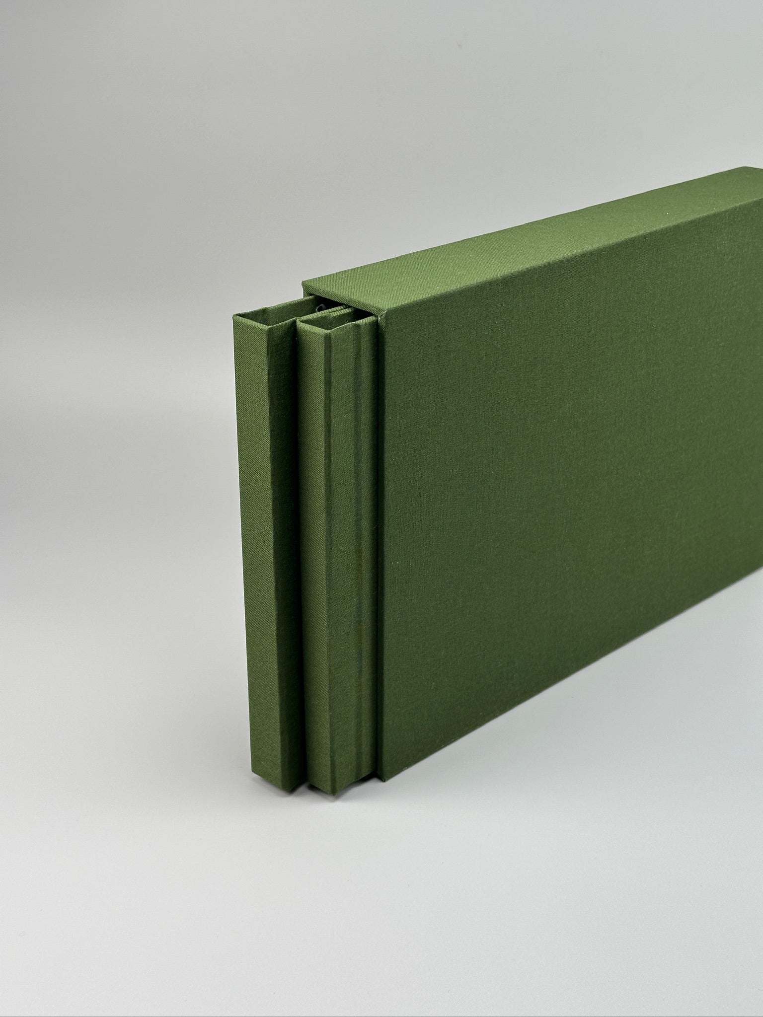 Estuche apaisado de lino verde oliva de canto, con el fotolibro entreabierto en el interior