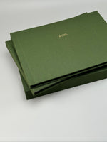 Fotolibro apaisado de lino verde oliva con nombre en dorado, apoyado sobre su caja a juego