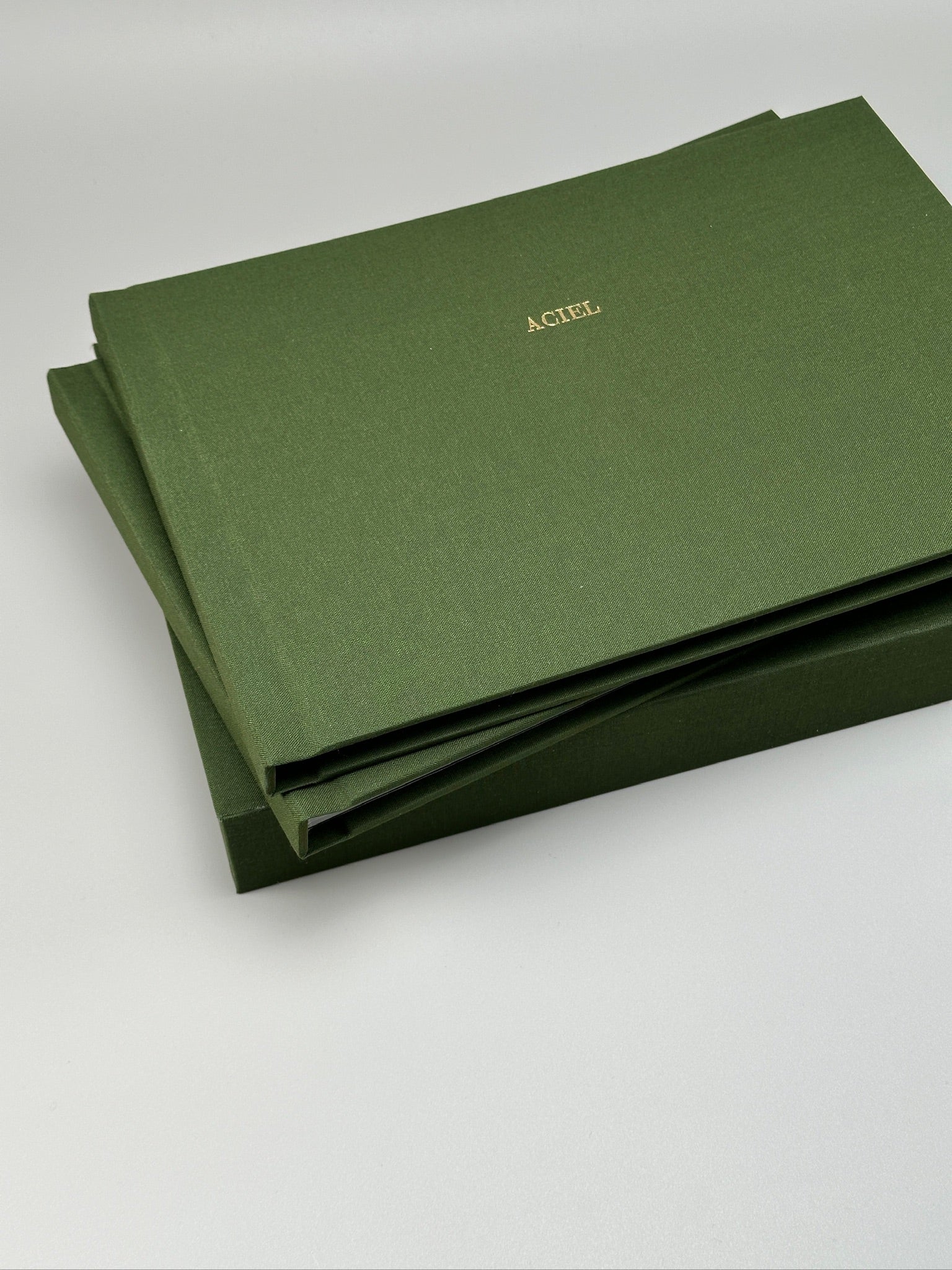 Fotolibro apaisado de lino verde oliva con nombre en dorado, apoyado sobre su caja a juego