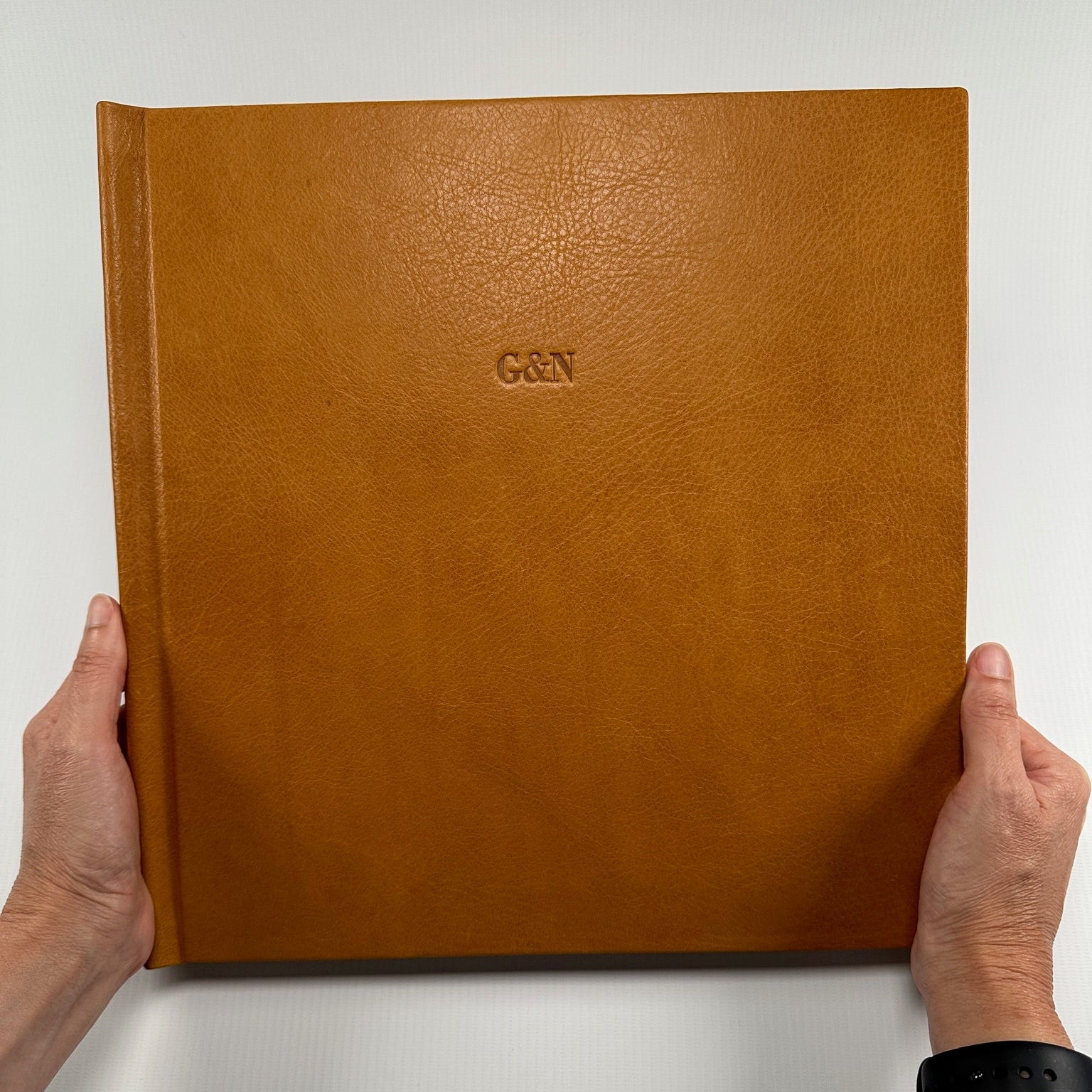 Manos sosteniendo un fotolibro cuadrado de cuero color cognac con las iniciales G&N grabadas en la tapa