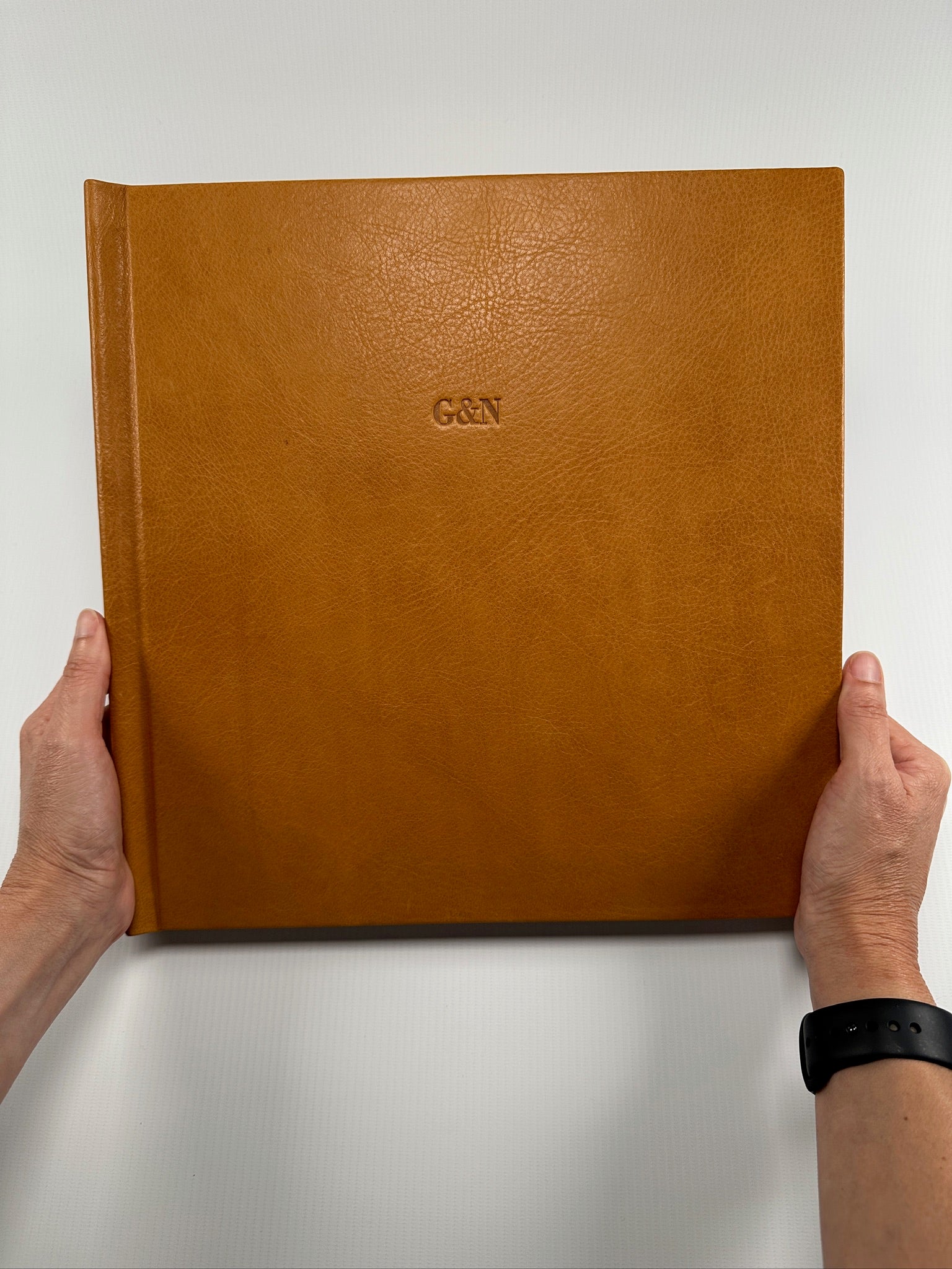 Manos sosteniendo un fotolibro cuadrado de tapa de cuero color caramelo con las iniciales G&N en relieve