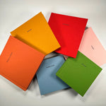 Variedad de Fotolibros Premium Layflat en distintos colores de lino (naranja, amarillo, rojo, rosa, verde y celeste), personalizados con el nombre del destino