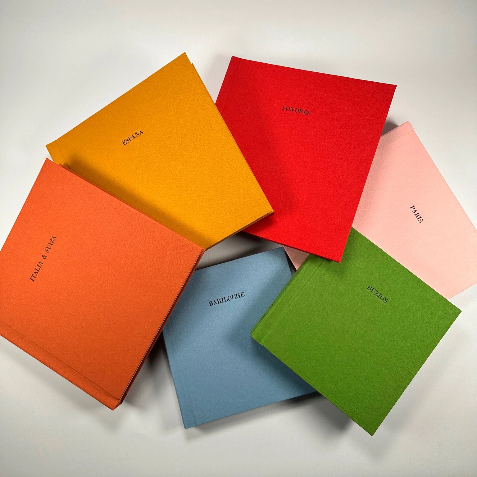 Variedad de Fotolibros Premium Layflat en distintos colores de lino (naranja, amarillo, rojo, rosa, verde y celeste), personalizados con el nombre del destino