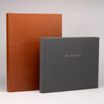 Dos libros de firmas premium: uno vertical de cuero color cognac y otro horizontal de lino gris con título dorado grabado